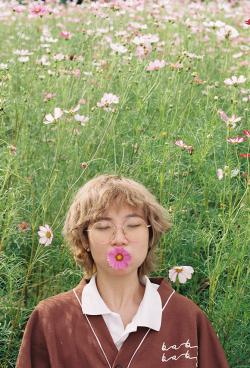 Eine Teenagerin steht vor einer schönen Blumenwiese. Sie hält eine Blume im Mund und hat dabei verschlossene Augen.