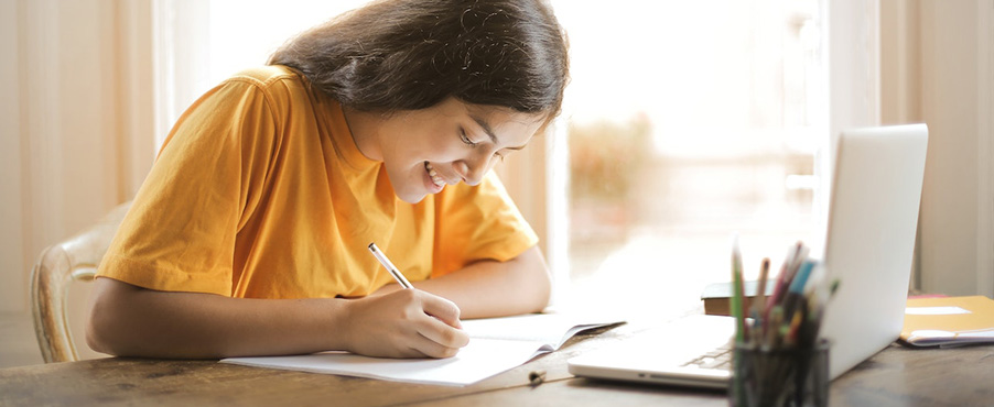 Eine Jugendliche sitzt an einem Schreibtisch und schreibt etwas auf. Auf dem Tisch stehen auch ein Laptop und ein Stiftebecher.