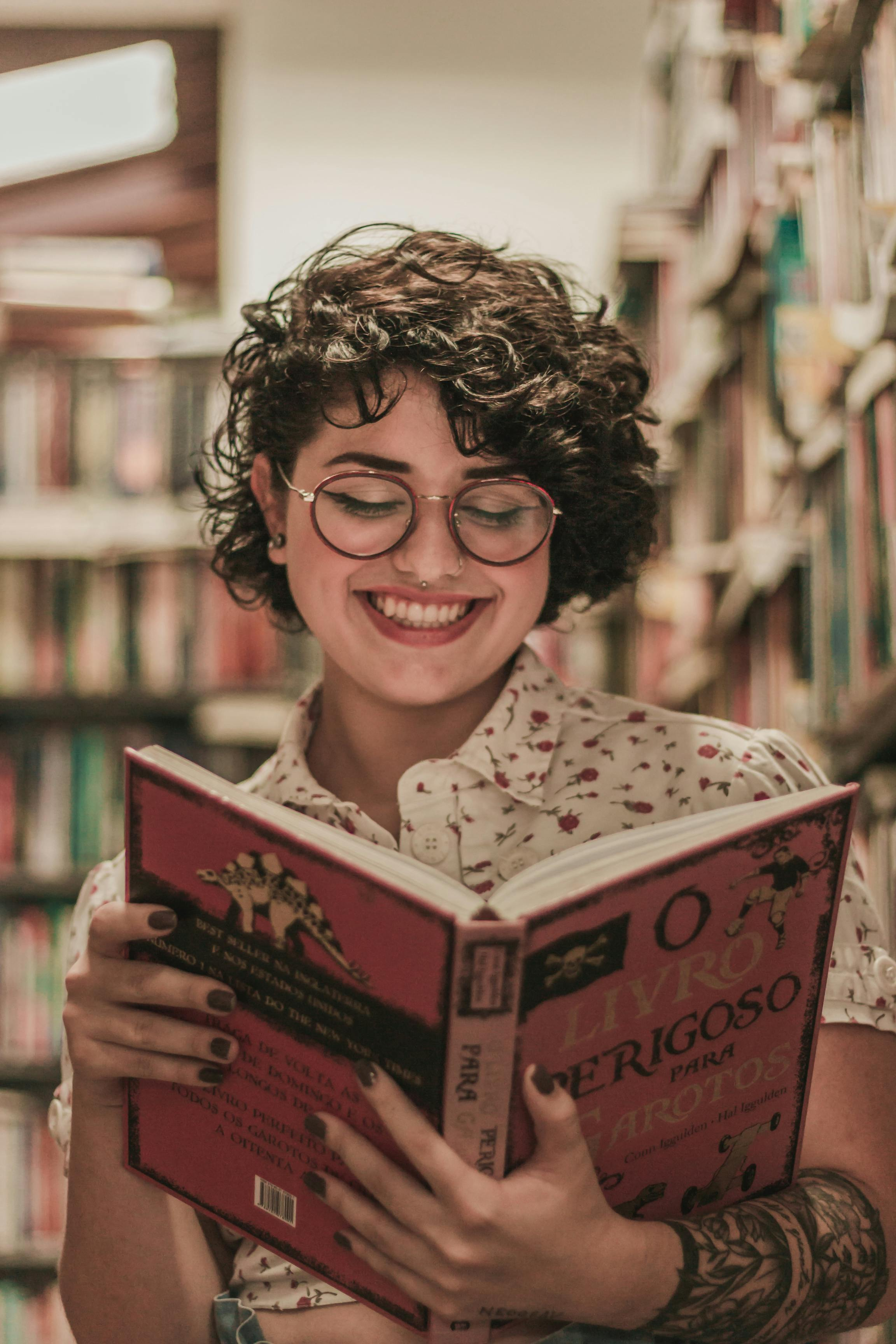 Eine junge Frau liest lächelnd in einem Buch. Sie steht vor einem Bücherregal.