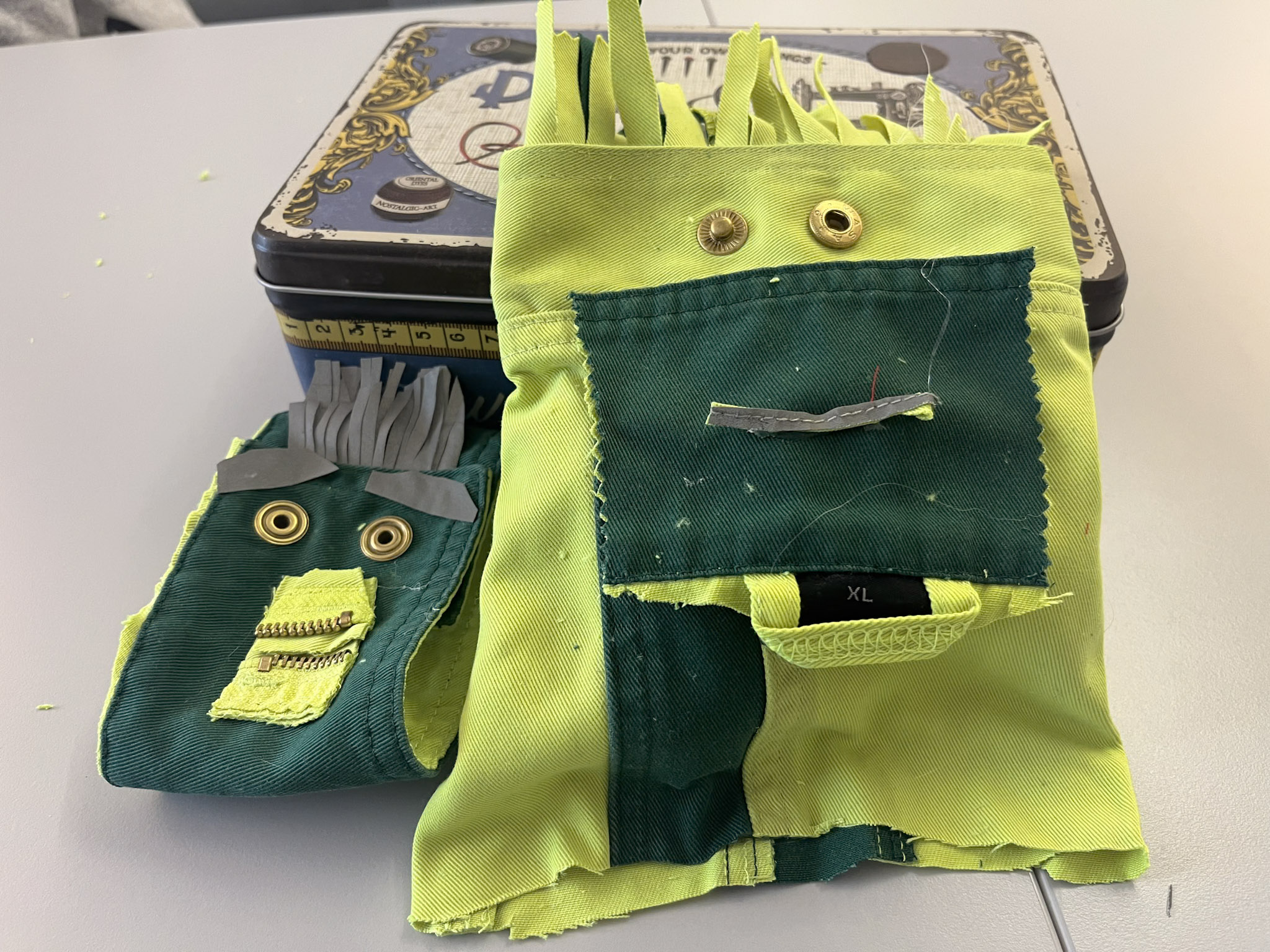 Zwei aus Stoffresten gebastelte Figuren liegen vor einer Metallbox: Eine kleine grün-gelbe Figur mit Reißverschluss-Mund und Knopfaugen sowie eine größere Figur aus gelbem und grünem Stoff mit Knopfaugen und Stofffransen