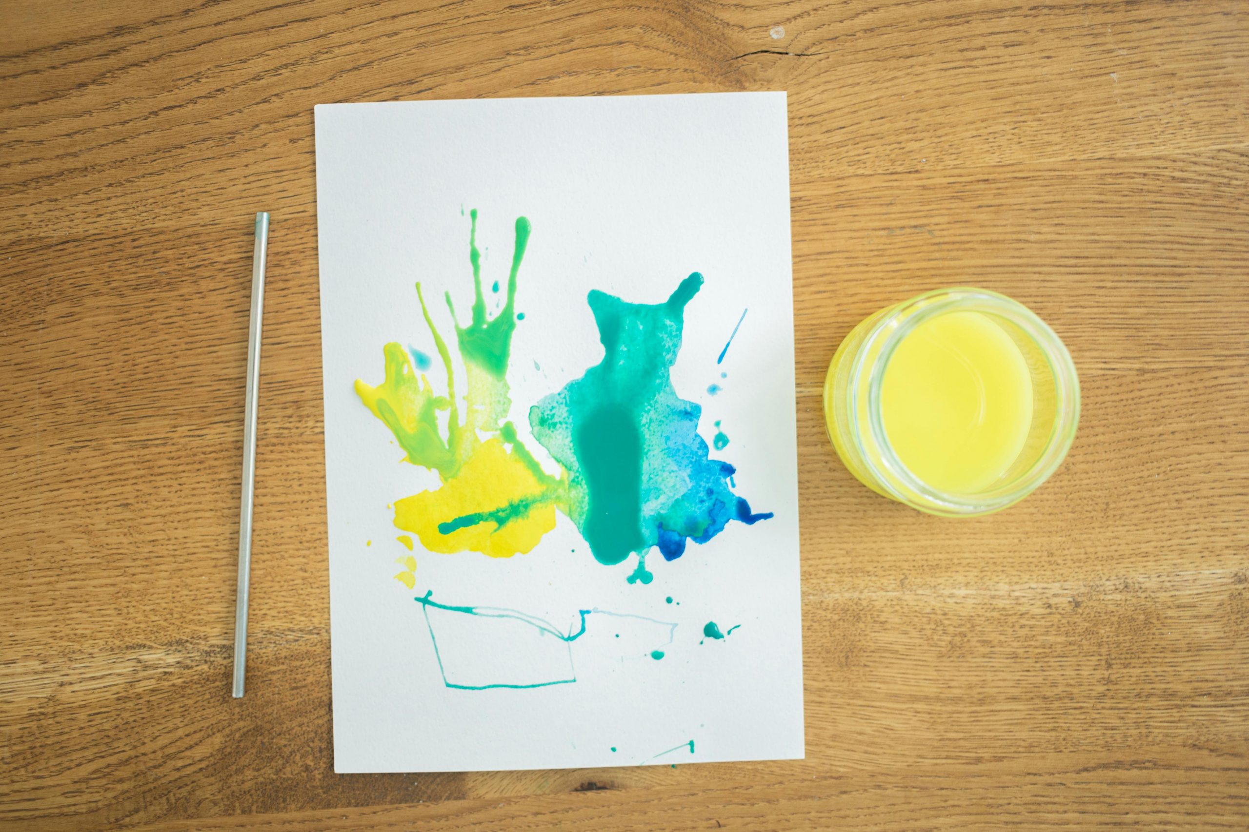 Auf dem Bild ist ein Blatt Papier und ein Töpfchen mit gelber Farbe zu sehen. Die Farbe wurde auf ein Blatt geklekst und mit einem Strohhalm in verschiedene Richtungen gepustet.