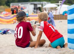 An einem Strand sitzen zwei Jungen mit dem Rücken zum Betrachter und lesen einen Comic.